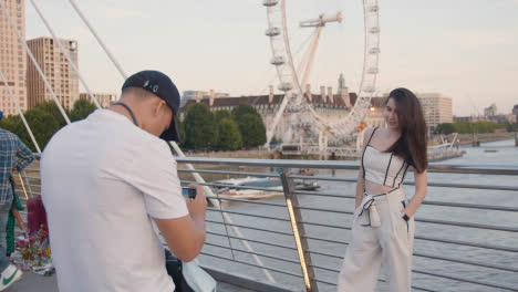 Tourist-Couple-Posing-For-Photo-On-Hungerford-Pedestrian-Bridge-With-London-Eye-In-Background-England-UK