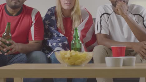Multi-Cultural-Friends-Supporting-USA-Team-Watching-Sports-Game-On-TV-Sitting-On-Sofa-At-Home-With-Flag