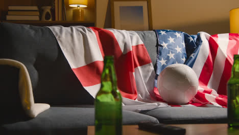 Sofa-In-Der-Lounge-Mit-Englischen-Und-Amerikanischen-Flaggen-Und-Ball,-Während-Fans-Fußballspiele-Im-Fernsehen-Sehen