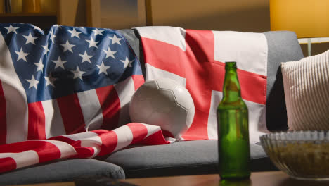 Sofa-In-Der-Lounge-Mit-Englischen-Und-Amerikanischen-Flaggen-Und-Ball,-Während-Fans-Fußballspiele-Im-Fernsehen-Verfolgen-3