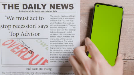 Zeitung-Mit-Schlagzeile-über-Rezession-Neben-Der-Hand,-Die-Das-Handy-Mit-Grünem-Bildschirm-Aufhebt