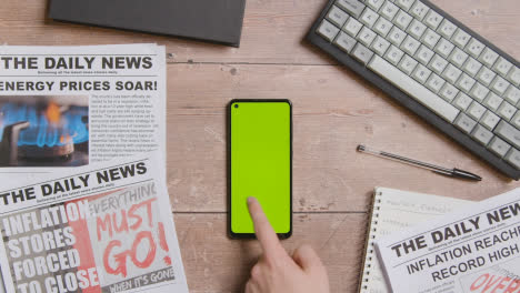 Overhead-Shot-Of-Table-With-Hand-Scrolling-Through-Green-Screen-Mobile-Phone-With-Newspaper-Stories-On-Inflation-And-Cost-Of-Living