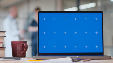 Laptop-Mit-Blauem-Bildschirm-Auf-Dem-Schreibtisch-Im-Krankenhaus-Mit-Arzt-Und-Personal-Im-Hintergrund