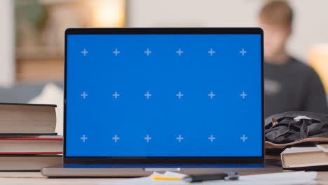 Blue-Screen-Laptop-on-Desk-At-Home-With-Student-Working-In-Background