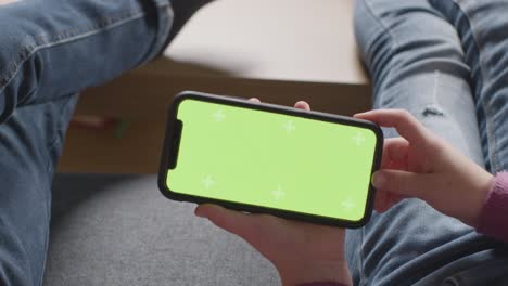 Close-Up-Of-Two-Children-Sitting-On-Sofa-At-Home-Looking-At-Green-Screen-Mobile-Phone-1