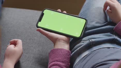 Close-Up-Of-Two-Children-Sitting-On-Sofa-At-Home-Looking-At-Green-Screen-Mobile-Phone-3