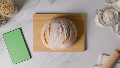 Overhead-Shot-Of-Freshly-Baked-Loaf-Of-Bread-Being-Picked-Up-From-Wooden-Board-With-Digital-Tablet
