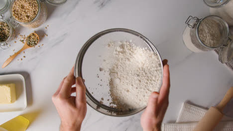 Overhead-Shot-Of-Flour-Being-Sieved-Onto-Marble-Work-Surface-For-Baking-With-Ingredients