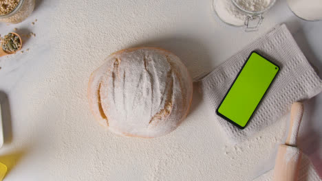 Überkopfaufnahme-Eines-Frisch-Gebackenen-Brotlaibs,-Der-Mit-Einem-Mobiltelefon-Mit-Grünem-Bildschirm-Auf-Eine-Marmorarbeitsfläche-Gelegt-Wird