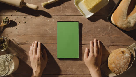 Overhead-Shot-Of-Green-Screen-Digital-Tablet-On-Wooden-Table-With-Freshly-Baked-Loaves-Of-Bread