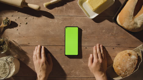 Overhead-Shot-Of-Green-Screen-Mobile-Phone-On-Wooden-Table-With-Freshly-Baked-Loaves-Of-Bread