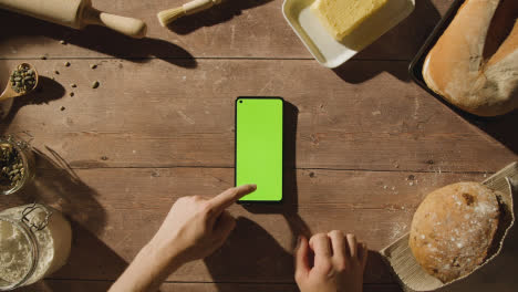 Overhead-Shot-Of-Person-Using-Green-Screen-Mobile-Phone-On-Wooden-Table-With-Freshly-Baked-Loaves-Of-Bread