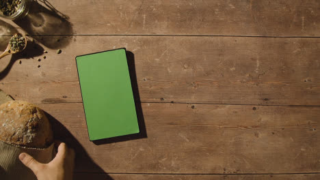 Overhead-Shot-Of-Person-Picking-Up-Loaf-Of-Bread-From-Wooden-Table-With-Green-Screen-Digital-Tablet