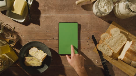 Überkopfaufnahme-Einer-Person,-Die-Frisch-Gebackenes-Brot-Mit-Butter-Auf-Einem-Holztisch-Mit-Einem-Digitalen-Tablet-Mit-Grünem-Bildschirm-Isst