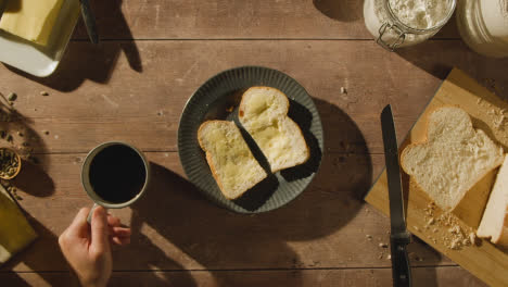 Overhead-Shot-Of-Person-Eating-Freshly-Baked-Bread-With-Butter-On-Wooden-Table-With-Hot-Drink