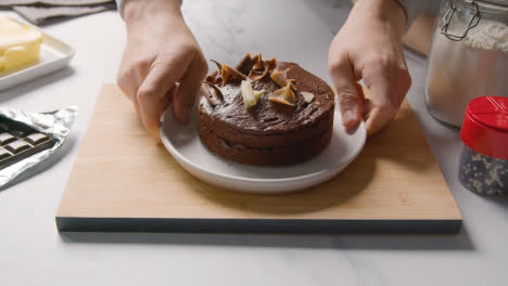 Person-Putting-Homemade-Chocolate-Cake-On-Plate-Onto-Kitchen-Work-Surface-With-Ingredients