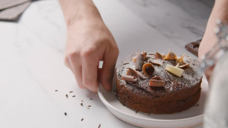 Person-Picking-Up-Homemade-Chocolate-Cake-On-Plate-On-Kitchen-Work-Surface-Behind-Ingredients