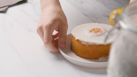 Person-Picking-Up-Homemade-Lemon-Drizzle-Cake-On-Plate-On-Kitchen-Work-Surface-Behind-Ingredients