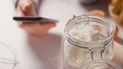 Close-Up-Of-Messy-Kitchen-Surface-With-Ingredients-And-Utensils-And-Person-Using-Mobile-Phone