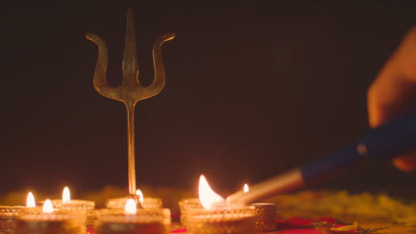 Hand-Lighting-Tea-Lights-Around-Metal-Trishula-Statue-Divine-Trident-Symbol-Of-Hinduism-On-Decorated-Table