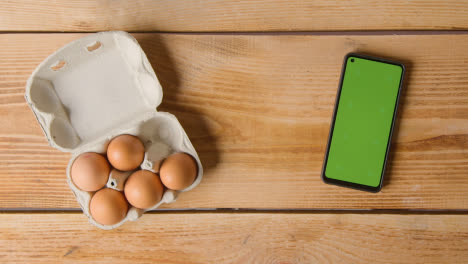 Fotografía-Cenital-De-Seis-Huevos-En-Una-Caja-De-Cartón-Que-Se-Abre-En-Una-Mesa-De-Madera-Con-Teléfono-Móvil