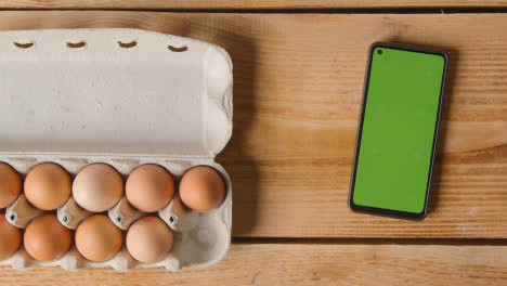 Fotografía-Cenital-De-Una-Persona-Que-Elige-Un-Huevo-De-Una-Caja-De-Cartón-En-Una-Mesa-De-Madera-Con-Teléfono-Móvil