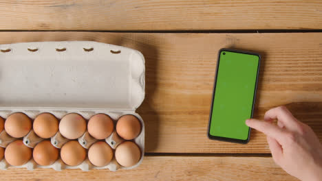 Fotografía-Cenital-De-La-Persona-Que-Sigue-La-Receta-En-Un-Teléfono-Móvil-De-Pantalla-Verde-Con-Una-Caja-De-Huevos-En-Una-Mesa-De-Madera