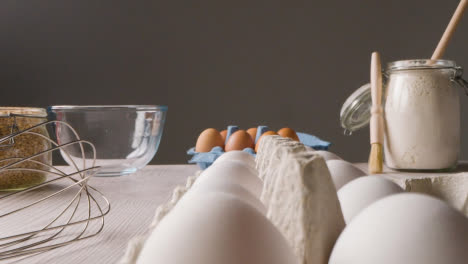 Studio-Shot-Of-Baking-Ingredients-And-Utensils-On-Kitchen-Worktop-With-Person-Picking-Up-Egg