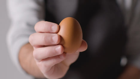 Close-Up-Studio-Shot-Of-Person-In-Apron-Holding-Up-Brown-Egg-To-Camera