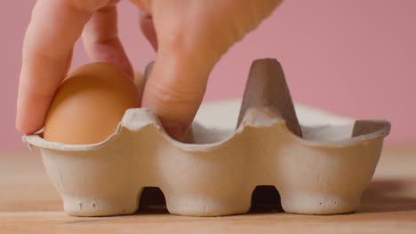 Studio-Shot-Of-Person-Taking-Brown-Eggs-From-Open-Cardboard-Box-Against-Pink-Background