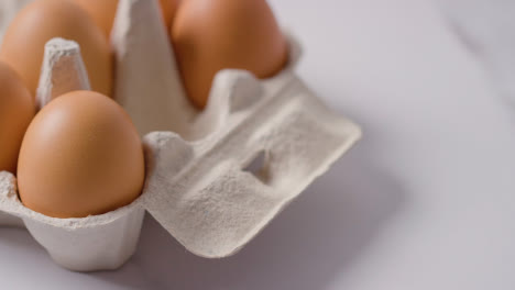 Studio-Shot-Of-Person-Putting-Egg-Into-Open-Cardboard-Box-On-Marble-Work-Surface-Background