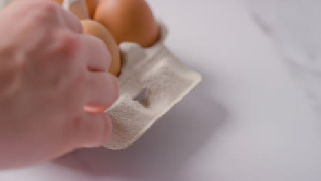 Studio-Shot-Of-Person-Putting-Egg-Into-Open-Cardboard-Box-On-Marble-Work-Surface-Background