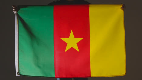 Studioaufnahme-Einer-Anonymen-Person-Oder-Eines-Sportfans,-Der-Die-Flagge-Kameruns-Vor-Schwarzem-Hintergrund-Hält