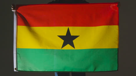 Studioaufnahme-Einer-Anonymen-Person-Oder-Eines-Sportfans,-Der-Die-Flagge-Von-Ghana-Vor-Schwarzem-Hintergrund-Hält