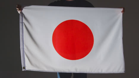 Studioaufnahme-Einer-Anonymen-Person-Oder-Eines-Sportfans,-Der-Die-Flagge-Japans-Vor-Schwarzem-Hintergrund-Hält