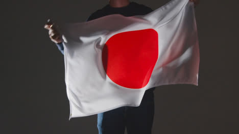 Studio-Shot-Of-Anonymous-Person-Or-Sports-Fan-Waving-Flag-Of-Japan-Against-Black-Background