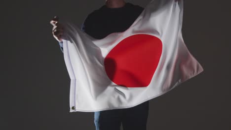 Studio-Shot-Of-Anonymous-Person-Or-Sports-Fan-Waving-Flag-Of-Japan-Against-Black-Background