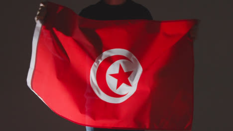 Studio-Shot-Of-Anonymous-Person-Or-Sports-Fan-Waving-Flag-Of-Tunisia-Against-Black-Background