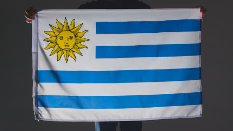 Studioaufnahme-Einer-Anonymen-Person-Oder-Eines-Sportfans,-Der-Die-Flagge-Von-Uruguay-Vor-Schwarzem-Hintergrund-Hält