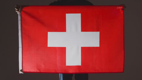 Studio-Shot-Of-Anonymous-Person-Or-Sports-Fan-Holding-Flag-Of-Switzerland-Against-Black-Background