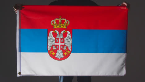 Studioaufnahme-Einer-Anonymen-Person-Oder-Eines-Sportfans,-Der-Die-Flagge-Serbiens-Vor-Schwarzem-Hintergrund-Hält