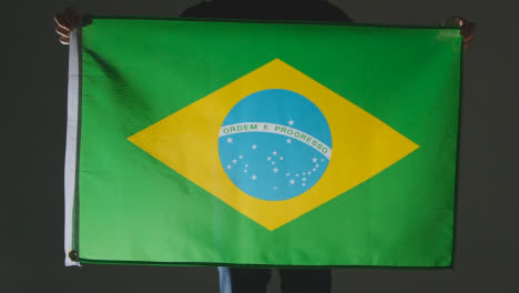 Studio-Shot-Of-Anonymous-Person-Or-Sports-Fan-Holding-Flag-Of-Brazil-Against-Black-Background