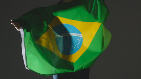 Studio-Shot-Of-Anonymous-Person-Or-Sports-Fan-Waving-Flag-Of-Brazil-Against-Black-Background