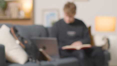 Defocused-Shot-Of-College-Student-Reading-Book-And-Working-On-Laptop-At-Home