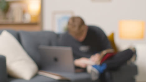 Defocused-Shot-Of-College-Student-Reading-Book-And-Working-On-Laptop-At-Home-1