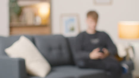 Defocused-Shot-Of-Young-Man-Sitting-On-Sofa-At-Home-Using-Mobile-Phone
