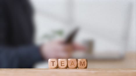 Business-Concept-Wooden-Letter-Cubes-Or-Dice-Spelling-Team-With-Office-Person-Using-Mobile-Phone-In-Background