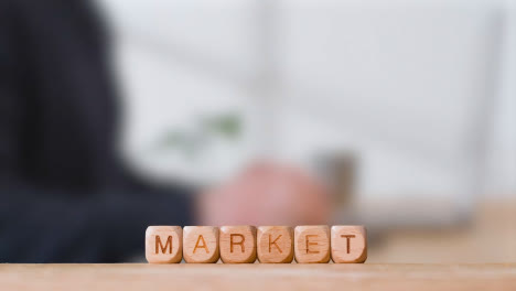 Business-Concept-Wooden-Letter-Cubes-Or-Dice-Spelling-Market-With-Office-Person-Using-Mobile-Phone-In-Background