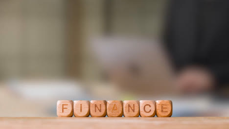Business-Concept-Wooden-Letter-Cubes-Or-Dice-Spelling-Finance-With-Person-Working-On-Laptop-In-Office-In-Background
