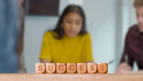 Business-Concept-Wooden-Letter-Cubes-Or-Dice-Spelling-Success-With-Office-Meeting-In-Background